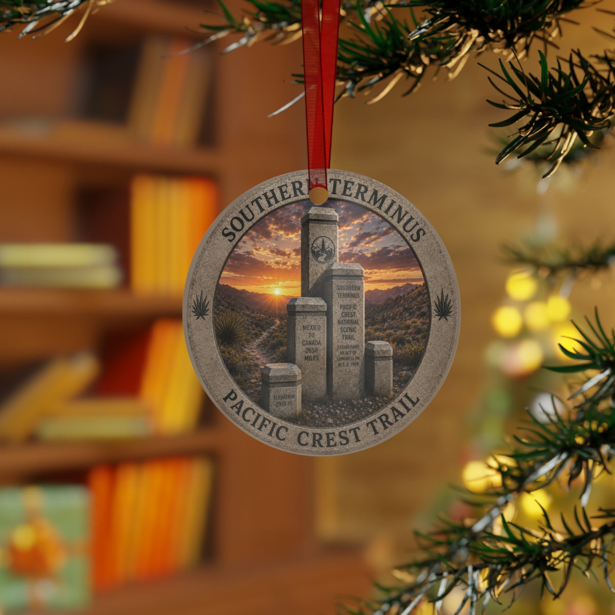 A round Christmas ornament hanging from a red ribbon on a Christmas tree branch. The ornament features an engraved design of the Southern Terminator and Pacific Crest Trail monument with a sunset sky in the center, set against a blurred background of a bookshelf and warm holiday lights.